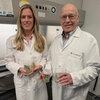  man and a woman in white lab coats hold containers with young green plants in a lab setting.